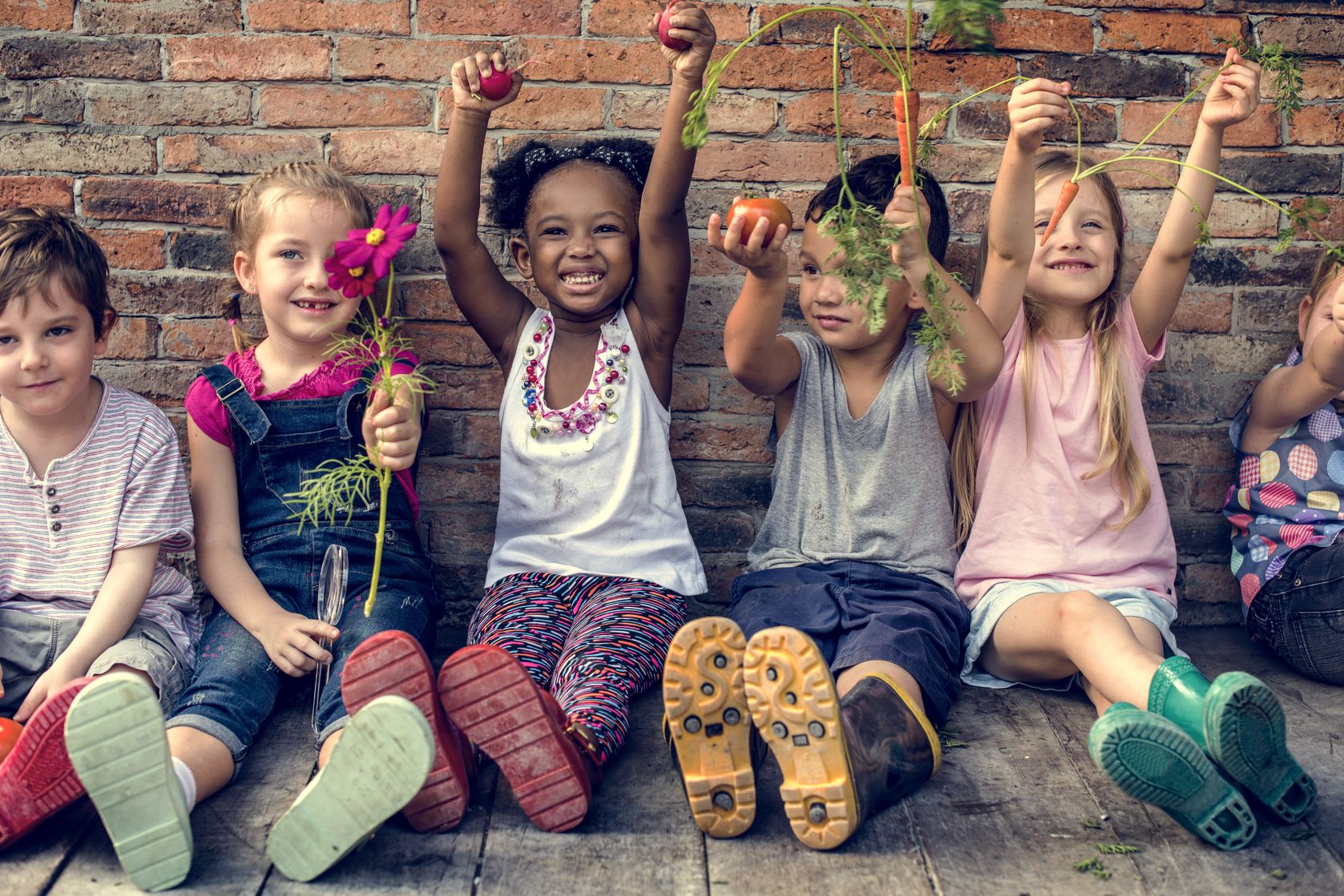 kids sitting in front of wall and holding vegetables and flowers 